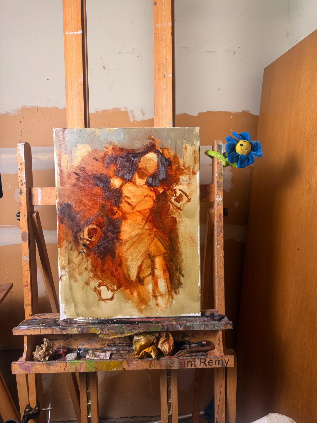 Work in progress on the easel — oil underpainting, studio, with a small blue flower clipped to the easel